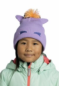 Gorro de punto púrpura con acentos en forma de orejas y pompón naranja. Presenta pestañas bordadas en azul. Llevado con una chaqueta verde menta.