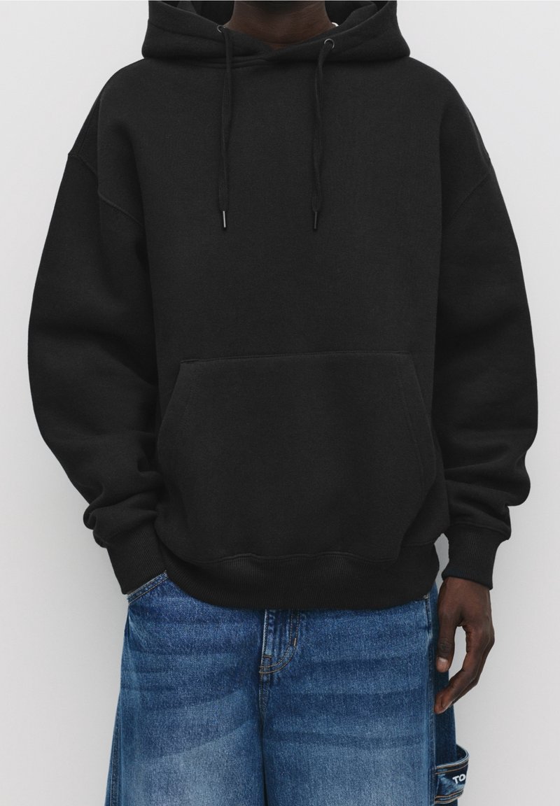 Homme portant un sweat à capuche noir uni avec poche avant et un jean en denim bleu, debout devant un fond blanc.