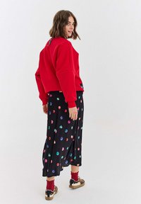 Červená zkrácená mikina, černá sukně s barevnými puntíky, doplněná vzorovanými teniskami. Viditelné detaily zahrnují žebrované manžety a lem.