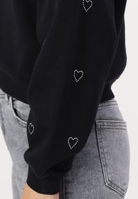 Manche de sweat-shirt noir avec petits motifs de cœurs en strass à côté de la poche et de la ceinture d'un jean gris clair.