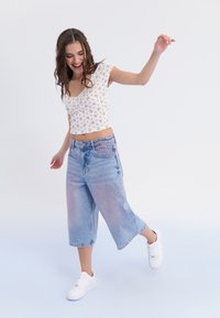 Cvjetni print na kratkom topu; svijetle traperice culottes sa izblijeđenim efektom; bijele tenisice; model u dinamičnoj pozi na jednostavnoj pozadini.