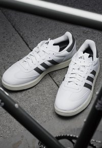 Białe skórzane sneakersy z trzema czarnymi paskami, teksturowaną gumową podeszwą i wyściełanym kołnierzem dla komfortu. Minimalistyczny design, klasyczny styl.