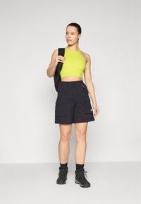 Svarta cargo-shorts med flera fickor, matchade med en smal, ljusgrön crop top. Tillbehör inkluderar en svart ryggsäck och mörka stövlar.
