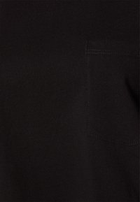 Tissu noir avec une petite poche carrée cousue sur le côté droit, montrant des détails de couture fins.