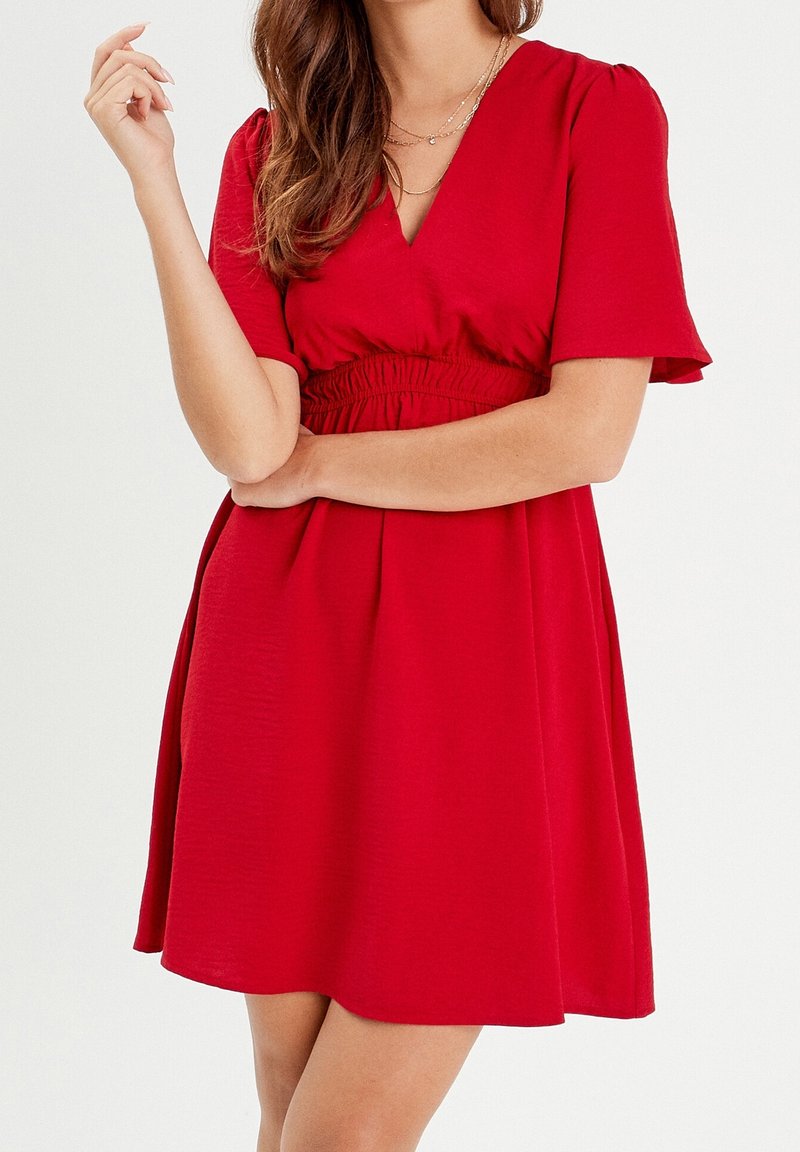 Femme portant une robe rouge à manches courtes avec un décolleté en V et une taille froncée, visible de la mi-cuisse aux épaules, avec la main près du visage.