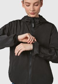 Black lightweight jacket with a zip front, a hood, elastic cuffs, and a smooth texture. Features brand logo on the zipper.