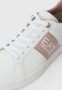 Beli usnjeni copati z roza in zlatimi detajli, ki vključujejo logotip, bele vezalke, gladko teksturo in gumijast podplat.