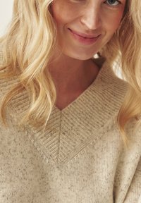 Beige kötött pulóver mély V-nyakkivágással, strukturált bordázással a nyakrésznél és puha, márványos anyagból készült. Természetes szálakból, laza szabású.