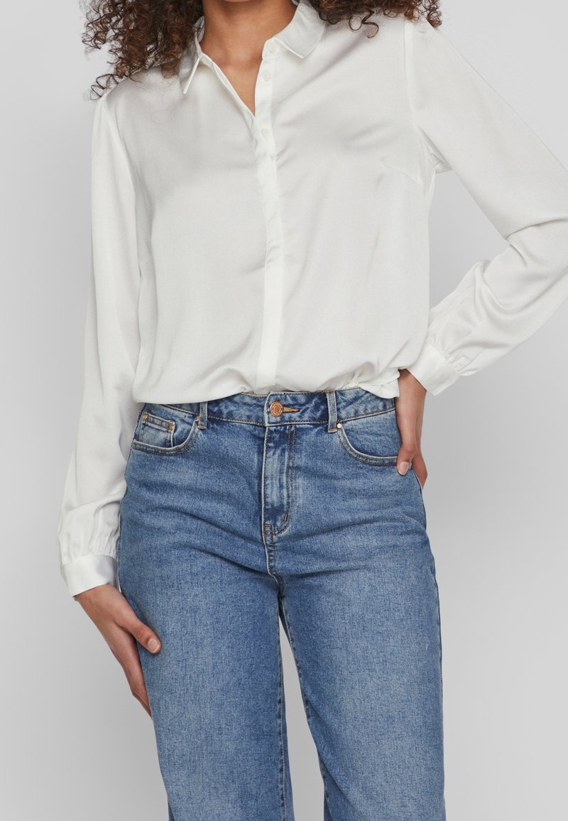 Femme portant une chemise blanche à manches longues rentrée dans un jean bleu taille haute, une main dans la poche et l'autre détendue le long du corps.