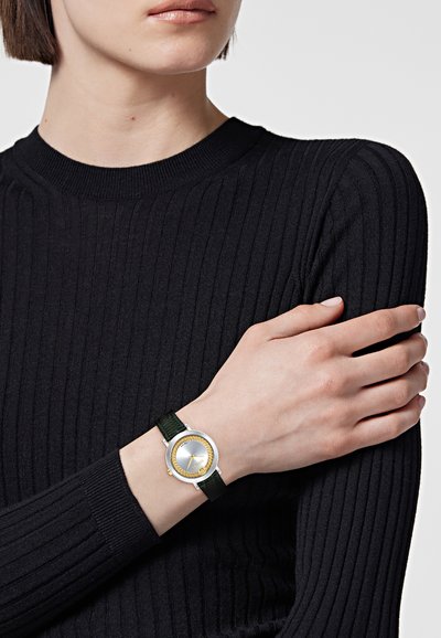 Personne portant un pull côtelé noir et une montre-bracelet avec un cadran rond argenté et doré et un bracelet en cuir vert foncé.