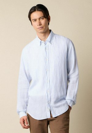 Junger Mann mit glatten dunklen Haaren trägt ein hellblau-weiß gestreiftes Hemd mit Knopfleiste und braune Hose, posiert vor beigem Hintergrund.