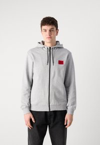 Šedá mikina na zip s přední kapsou a kapucí se stahovací šňůrkou. Na hrudi má červený obdélníkový logo. Nosit s černými kalhotami.