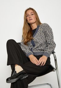 Štrikovaný kardigán v modro-sivej farbe s textúrovaným vzorom, výstrih do V a predným zapínaním na gombíky. Skombinovaný s čiernymi širokými nohavicami a mokasínami.