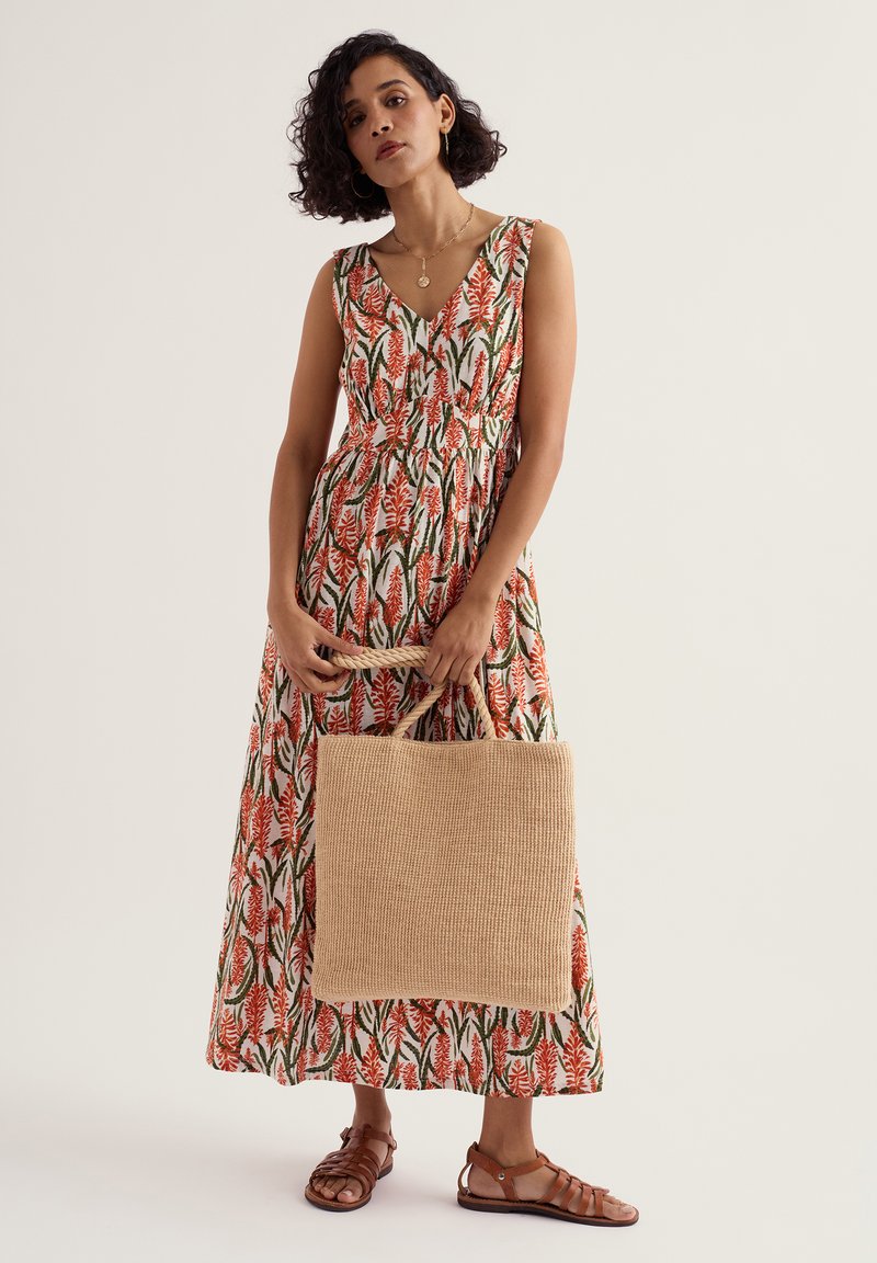 Femme portant une robe longue sans manches à fleurs, tenant un sac fourre-tout beige tissé, et des sandales plates marron sur fond uni.