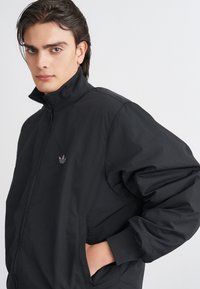 Crna vjetrobran jakna s visokim ovratnikom, prednjim zatvaračem i rebrastim manžetama. Sadrži mali Adidas logo na lijevoj strani prsnog dijela. Glatka tekstura.