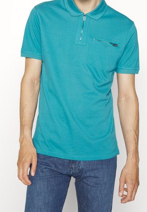Mann trägt ein türkisfarbenes kurzärmeliges Polo mit Frontreißverschluss und Brusttasche mit Reißverschluss, kombiniert mit blauen Jeans, vor weißem Hintergrund.