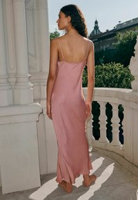 Abito in satin rosa con spalline sottili, silhouette aderente e orlo fluente. Texture liscia; dettagli minimi, indossato a piedi nudi su un balcone.