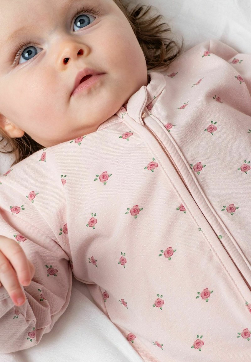 Bébé aux yeux bleus portant une grenouillère fleurie rose pâle, allongé et regardant légèrement vers le haut et sur le côté.