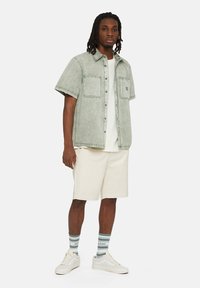 Camicia verde chiaro a maniche lunghe con due tasche sul petto, abbinata a una maglietta bianca, pantaloni beige e scarpe da ginnastica bianche con calzini a righe verdi.