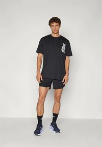 Giovane uomo in piedi, indossa pantaloni corti sportivi neri, una maglietta nera "One More Mile", calze nere Adidas e scarpe da corsa nere.