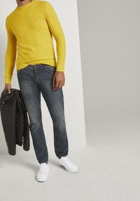 Żółty sweter z długim rękawem o prążkowanej fakturze, wyblakłe niebieskie dżinsy, białe trampki i czarna skórzana kurtka trzymana w lewej ręce.