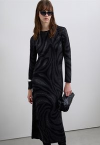 Abito nero a maniche lunghe con motivo metallico vorticoso, completato da una pochette nera e occhiali da sole. Silhouette liscia e aderente.