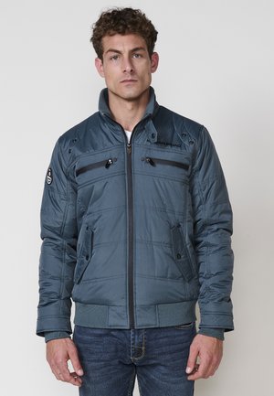 Chaqueta acolchada gris con un cuello alto, bolsillos con cremallera y puños elásticos. Presenta detalles cosidos y un logotipo en el hombro.