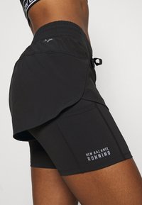 Närbild av en person som bär svarta, lager-på-lager löpshorts med texten "New Balance Running" på det inre lagret mot en enfärgad bakgrund.