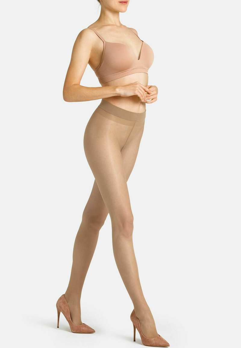 Beige sømløs bralette parret med gennemsigtige nude strømpebukser. Højtaljet med en glat tekstur. Modellen bærer pink ruskind stiletto pumps.