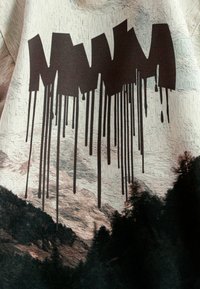 Texto gráfico negro "MMMM" con efecto de goteo sobre una tela clara y texturizada que presenta un paisaje montañoso en tonos tierra apagados.