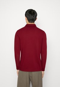 Poloko s dlouhými rukávy v barvě vínové s jemnou texturou, špičatým límcem a ribkovanými manžetami; zadní pohled ukazuje čisté linie a jednoduchý design.