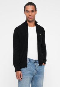 Juodas zip-up megztinis su raštuotu apykakle ir rankogaliais, dėvimas ant baltos marškinėlių, derinamas su šviesiai mėlynomis džinsinėmis kelnėmis. Matomas Lacoste logotipas.