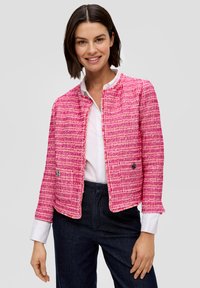 Vaaleanpunainen tweed-takki, jossa on pyöreä kaulus, kaksi etutaskua ja nypynnyt reuna. Päällä valkoinen paita ja tummat housut.