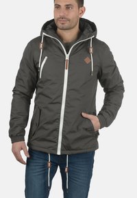 Donkergrijze hooded jack met een witte rits en bruine accenten. Voorzien van elastische manchetten en twee zijzakken. Gemaakt van lichtgewicht stof.