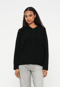 Svart v-hals genser i tykk strikk med en avslappet passform og lange ermer. Myk tekstur, ribbestrikkede mansjetter og kant. Kombinert med lysegrå jeans.