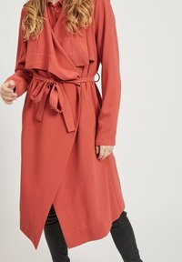 Płaszcz trench w kolorze rdzy z konstrukcją typu wrap, długimi rękawami, paskiem w talii i dwiema przednimi kieszeniami, wykonany z gładkiego materiału.
