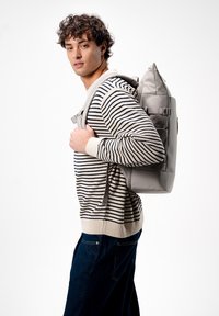 Zaino grigio con una superficie testurizzata e spallacci regolabili, indossato a tracolla. Maglione a righe, pantaloni scuri, sfondo minimale.