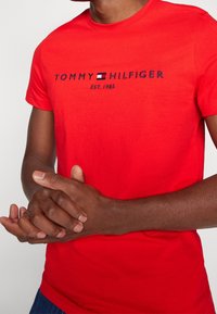 Rød bomulds T-shirt med broderet "TOMMY HILFIGER" logo og "EST. 1985" tekst. Korte ærmer, rund hals, slim fit.