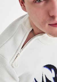 Κρεμ ζακέτα με φερμουάρ στο γιακά, με υφή υφάσματος και τολμηρό μαύρο και μπλε γραφικό σχέδιο στο μπροστινό μέρος.