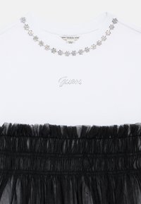 Biały top Guess z naszyjnikiem w kształcie kwiatów z kryształków na dekolcie i czarną marszczoną tiulową nakładką poniżej piersi.