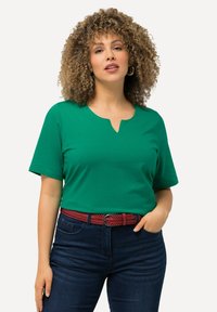 Top verde de manga corta con escote en forma de V, combinado con jeans azul oscuro y un cinturón trenzado rojo, resaltando una textura de tela suave.