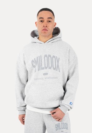 Ung mand iført lysegrå hættetrøje og joggingbukser med "SMILODOX Essential Sportswear" logo, stående mod en hvid baggrund.