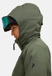 Person, der eine olivgrüne Kapuzenjacke und schwarze Skibrillen mit blau-grünen, reflexionsfähigen Gläsern trägt, nach links blickend.