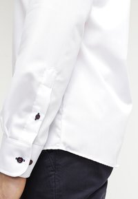 White dress shirt with long sleeves, featuring blue and red button accents on the cuffs. Smooth, textured fabric, and curved hem.