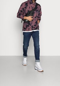 Sudadera morada con tie-dye, bolsa cruzada negra, jeans de mezclilla azules y zapatillas blancas de caña alta con suelas marrones. Los jeans remangados ofrecen un aspecto casual.
