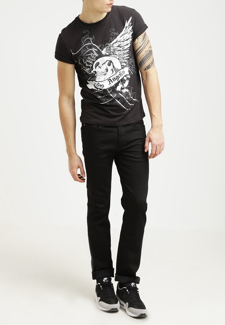 T-shirt graphique noir avec un motif de crâne et d'ailes, associé à un jean noir et des baskets noires, sur un fond neutre.
