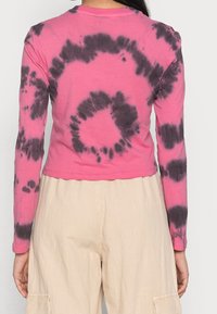 Pink long-sleeve shirt with black tie-dye patterns, cropped design, soft fabric, and round neck. Paired with beige trousers.