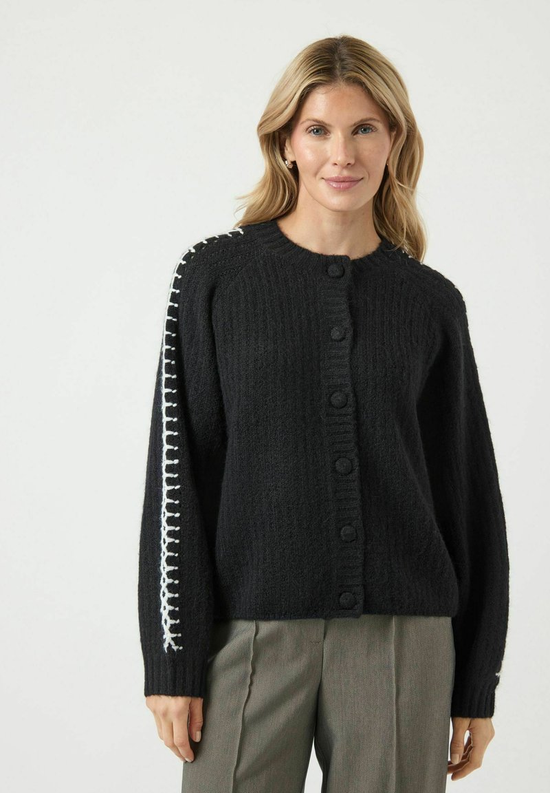 Svart stickad cardigan med rund halsringning, knappstängning och vit söm längs vänster ärm. Har en texturerad yta.