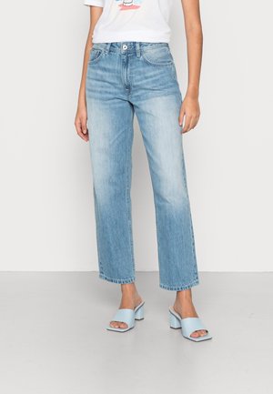 Nainen pukeutuneena vaaleansiniset, korkeavyötäröiset suoralahkeiset farkut, vaaleansiniset avokkaat paksulla koron ja valkoinen graafinen t-paita.