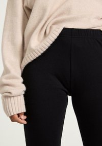 Beige neulepaita ribbikrauluilla, mustien legginssien päällä, joissa on joustava vyötärö, korostaen pehmeää, rakenteellista kangasta.